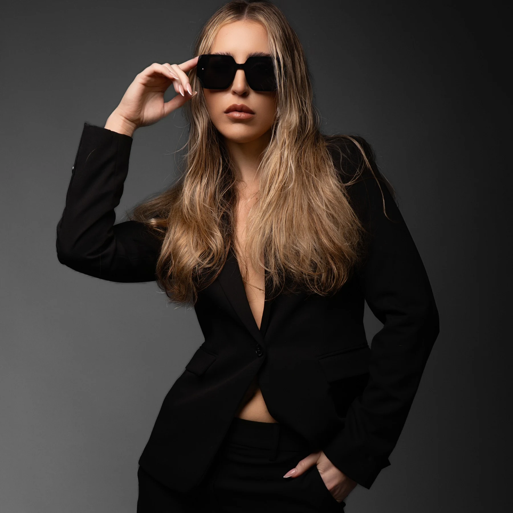 Editorial portrait in a black blazer and sunglasses against a dark background